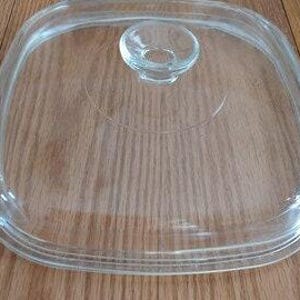 May include: A clear, square-shaped glass lid featuring rounded corners and a centered, circular glass knob handle. The lid has a stepped rim design that allows it to fit securely onto a corresponding square container or cookware piece.