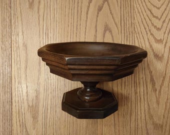 May include: A dark brown, wooden, hexagonal bowl-shaped shelf with a pedestal base. The shelf has a tiered design and is mounted on a wooden wall with a natural wood grain pattern. The shelf is likely for decorative purposes.