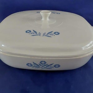 May include: A white, rectangular casserole dish with a matching lid. The dish and lid feature a delicate blue floral pattern. The lid has a white handle. The dish is on a blue surface.