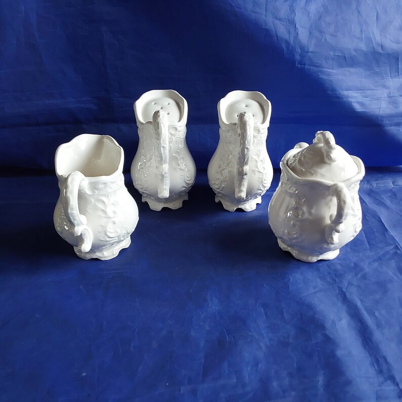 May include: Set of four white ceramic sugar and creamer containers with ornate floral designs. The containers have a vintage look and are perfect for a tea party or special occasion.