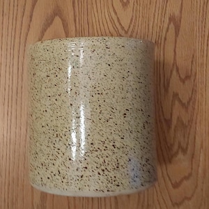 May include: A cylindrical ceramic vase with a speckled beige and black pattern. The vase has a smooth, glossy finish and a simple, modern design. The vase is approximately 6 inches tall.