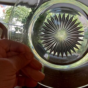 May include: Clear glass plate with a starburst design etched into the center. The plate is held up, showing the intricate pattern and the light passing through the glass. The plate's rim is visible, and the background is blurred.