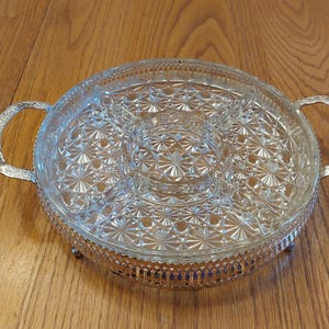 May include: A clear glass serving tray with a starburst pattern, divided into sections. It has a silver-colored metal base and handles. The tray is designed for serving appetizers or snacks.