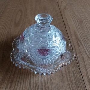 May include: A clear, cut-glass butter dish with a domed lid and a scalloped base. The dish features intricate geometric patterns and small pink accents. The lid has a knob on top for easy handling. The dish is on a wooden surface.