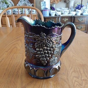 May include: An iridescent purple glass pitcher with a decorative grape and leaf design. The pitcher has a ruffled rim, a curved handle, and a wide base. The pitcher is sitting on a wooden table.