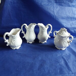 May include: A set of four white ceramic creamer and sugar containers with intricate floral designs. The containers have a vintage look and are perfect for serving coffee or tea.