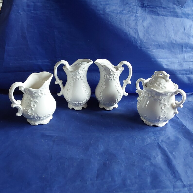 May include: A set of four white ceramic creamer and sugar containers with intricate floral designs. The containers have a vintage look and are perfect for serving coffee or tea.
