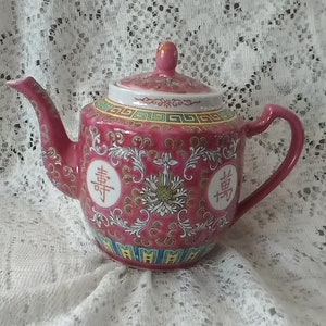 May include: A pink porcelain teapot with gold accents and Chinese characters. The teapot has a floral design and a lid with a knob.