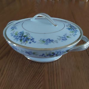 May include: A white porcelain serving dish with a lid, adorned with blue floral patterns and gold trim. The dish has two handles and a handle on the lid. A classic piece for serving food.