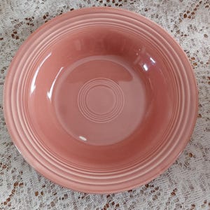 May include: A round, pink ceramic bowl with concentric rings around the rim and a central circle. The bowl is set against a white lace tablecloth. This is a piece of vintage tableware.