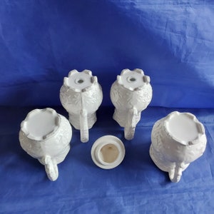 May include: Set of four white ceramic creamer or sugar containers with intricate floral designs. The containers have a round body and a spout. There is also a separate lid.