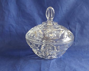 May include: A clear, cut-glass candy dish with a lid. The dish has a round, bulbous shape with a starburst pattern on the lid and sides. The lid has a loop handle. The dish is on a blue background.