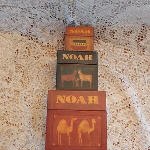 May include: A set of three stacked wooden boxes with the word "NOAH" printed on each box. The boxes are decorated with images of a boat, animals, and camels.