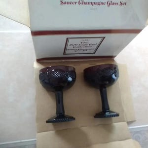 May include: A set of two dark red stemmed champagne glasses with a decorative pattern on the bowl. The glasses are displayed in an open box with text that reads "Saucer Champagne Glass Set" and "The 1870 Cape Cod Collection".