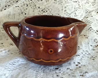 May include: A brown ceramic creamer with a scalloped rim and a floral design. The creamer has a handle and a spout.