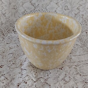 May include: A small, round, yellow ceramic bowl with a white speckled pattern. The bowl has a smooth, glossy finish and a slightly flared rim. The bowl is sitting on a white lace tablecloth.