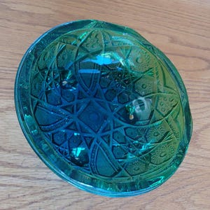 May include: A teal-colored, glass bowl with a detailed, geometric pattern. The bowl has a round shape with a slightly raised rim. The intricate design features star and diamond shapes. The bowl is sitting on a wooden surface.