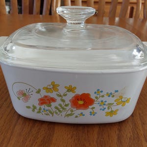 May include: White casserole dish with a clear glass lid and handle. The dish is decorated with a floral pattern featuring orange, yellow, and blue flowers. The dish has two side handles.