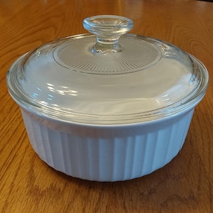 May include: A white, round casserole dish with a clear glass lid. The dish has vertical ribbed sides and a clear glass knob on top. The dish is sitting on a wooden table.