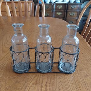May include: Three clear glass bottles in a black wire cage holder. The bottles are empty and the cage is made of a wire mesh.