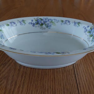 May include: Oval serving bowl with a white base, adorned with blue floral patterns and gold trim. The bowl has handles on each side and a gold rim. A decorative piece for serving food.