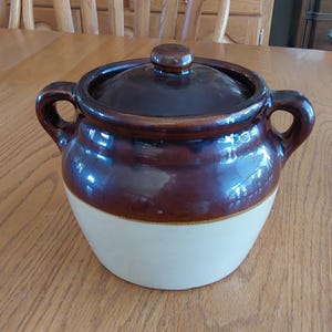 May include: A two-handled ceramic bean pot with a glossy dark brown glazed upper section and a matte cream-colored lower base. The pot features a matching dark brown lid with a small knob handle centered on top and two curved side handles.