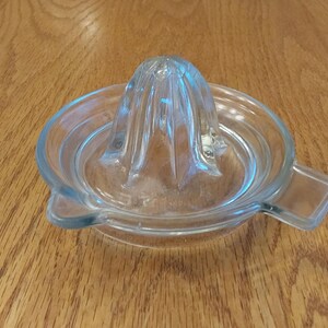 May include: A clear glass citrus juicer with a reamer and a bowl. The juicer has a ridged cone-shaped reamer in the center and a spout on the side. The juicer is designed for squeezing juice from citrus fruits.