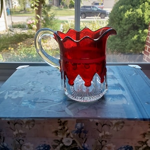 May include: A clear glass pitcher with a red overlay design. The pitcher has a handle and a decorative, scalloped rim. The red design drips down the sides, and the base is cut glass. The pitcher is sitting on a floral patterned surface.