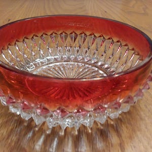 May include: A clear glass bowl with a ruby red rim and base. The bowl has a detailed, cut-glass design with a starburst pattern on the bottom. The bowl sits on a wooden surface.
