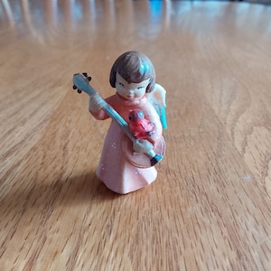 May include: A small figurine of an angel playing a banjo with a red bird perched on the instrument. The angel is wearing a pink dress with a blue halo and has brown hair.