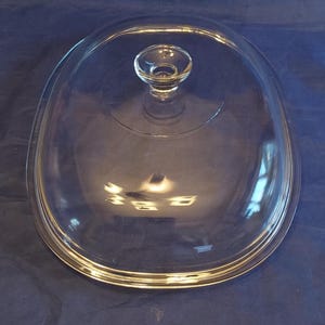 May include: Clear glass oval-shaped lid with a small, round handle. The lid is designed for cookware and is made of transparent glass, allowing visibility of the contents. The handle is also clear glass.