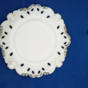 May include: White milk glass plate with a scalloped edge and a delicate floral design. The plate has a raised rim and a center well.