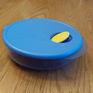 May include: A blue plastic food storage container with a matching lid. The lid has a yellow tab for easy opening. The container is on a wooden surface.