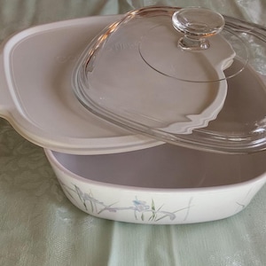 May include: A white casserole dish with a matching lid and a clear glass lid. The casserole dish has a floral design and is on a light green surface. The clear glass lid has a knob handle.