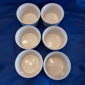 May include: Six white ceramic ramekins with a ribbed exterior and a smooth interior. The ramekins are arranged in two rows of three on a blue surface. These small dishes are ideal for baking individual portions.