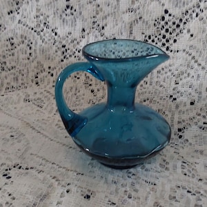 May include: A small, teal-colored glass pitcher with a handle and a spout. The pitcher has a rounded base and a textured surface. It is likely a decorative item or a small serving piece.