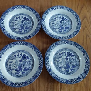 May include: Four blue and white ceramic plates with a classic Willow pattern. Each plate features a central landscape scene surrounded by a detailed border. The plates are arranged on a wooden surface.