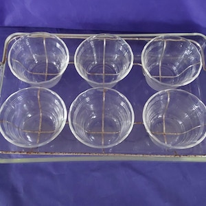 May include: A vintage serving set featuring six clear glass bowls held in a metal wire frame, set on a rectangular glass tray. The tray has handles on each side. The bowls are approximately 3 inches in diameter.