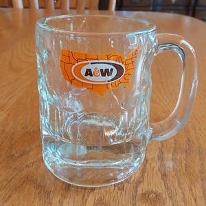 May include: A clear glass mug with a handle, featuring a sticker of the A&W logo and a map of the United States. The mug has a textured surface and is sitting on a wooden table.