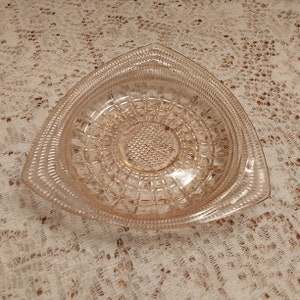 May include: A vintage pink glass triangular dish with a raised, textured design. The dish has a scalloped edge and a center circle with a smaller, raised, textured design.