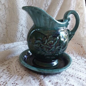 May include: A green ceramic pitcher with a floral design and a matching saucer. The pitcher has a wide mouth and a handle.