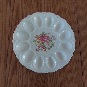 May include: Off-white ceramic egg serving platter with 12 egg-shaped indentations. The center features a floral design with pink, yellow, and red roses. The platter has a scalloped edge and is set on a wooden surface.