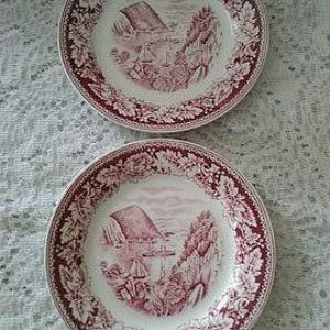 May include: Two matching ceramic plates with a red and white design. Each plate features a landscape scene with a building, trees, and a boat. The plates have a decorative border with a floral pattern.