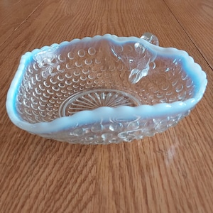 May include: Clear glass bowl with a light blue opalescent rim and a textured surface. The bowl has a handle and a decorative pattern on the base. The bowl is sitting on a wooden surface.