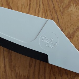 Op de afbeelding: Een witte Pampered Chef pizzasnijder met een zwarte snijkant. De snijder heeft een driehoekige vorm met een afgeronde hoek en het Pampered Chef logo.