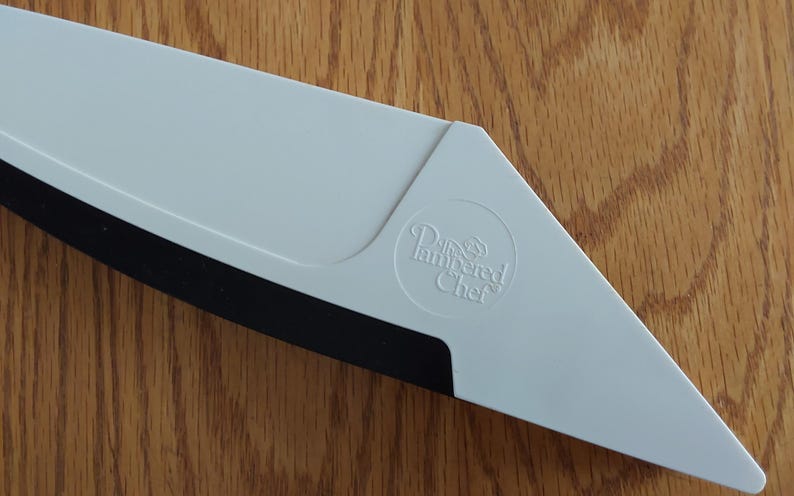 Op de afbeelding: Een witte Pampered Chef pizzasnijder met een zwarte snijkant. De snijder heeft een driehoekige vorm met een afgeronde hoek en het Pampered Chef logo.