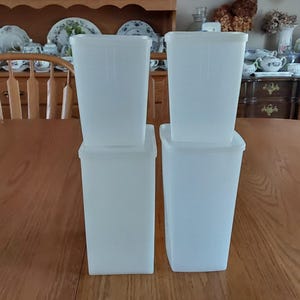 May include: Four translucent, rectangular food storage containers with lids are stacked on a wooden table. The containers are a light, off-white color and appear to be made of plastic. The background includes a wooden hutch and dining room furniture.