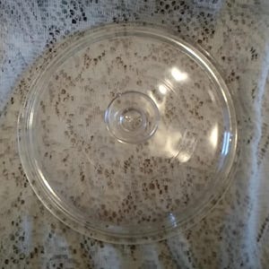 May include: A clear, round glass lid with a small knob in the center. The lid has a slightly raised rim and is designed to fit on a pot or pan. The glass is transparent, allowing visibility of the contents inside the cookware.