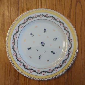 May include: A white ceramic plate with a yellow and gold rim, decorated with a floral pattern. The plate features a border of red and green leaves and small blue flowers. The center has scattered blue floral accents.