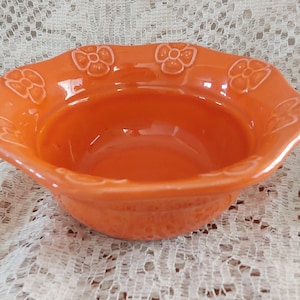 May include: An orange ceramic bowl with a scalloped rim and embossed floral designs. The bowl has a glossy finish and a shallow, rounded interior. The bowl is likely for serving or display.